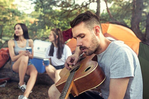 Party, camping of men and women group at forest. relaxing, singing a song against green grass.