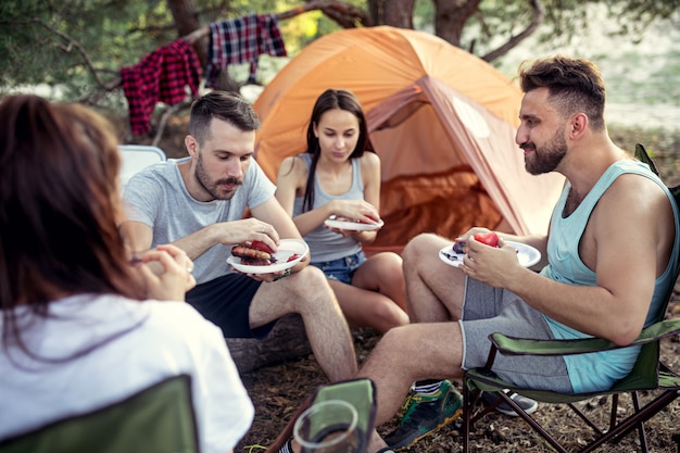 숲에서 남성과 여성 그룹의 파티, 캠핑. 그들은 편안하고 바베큐를 먹는