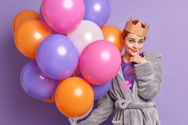 Party girl looks confident at front enjoys birthday celebration dressed in domestic outfit holds inflated colorful balloons poses indoor over purple wall