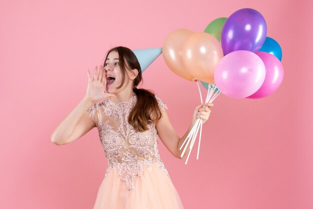 party girl with party cap holding balloons hailing someone on pink