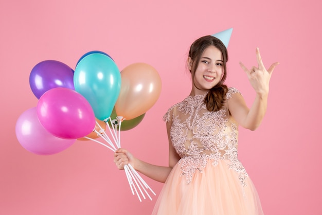 party girl with party cap holding balloons making rock sign on pink