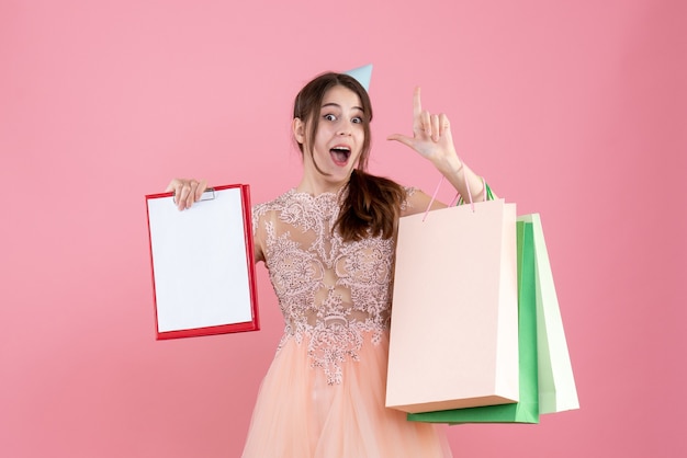 party girl with party cap holding documents and shopping bags pointing with finger up on pink
