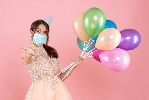 ピンクのカラフルな風船を保持しているパーティーキャップと医療マスクを持つパーティーの女の子