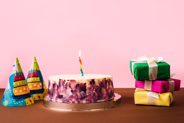 Party hat; birthday cake and stack of gift boxes on desk against pink background