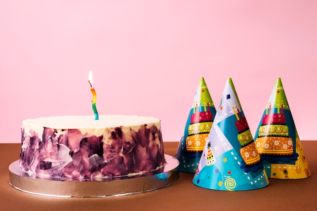 Party hats and cake with lighted candle on desk against pink backdrop