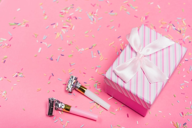 Party horns and closed gift box with colorful sprinkle on pink background