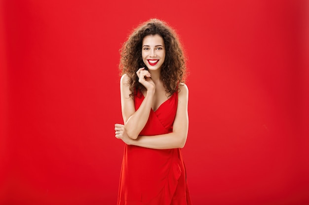 赤い背景の上の情熱的な魅力的なヨーロッパの女性は、巻き毛の髪型がかわいい笑顔で、臆病で愚かな髪の毛で遊んで、彼女が尊敬する人と話しているエレガントなドレスを着ています。