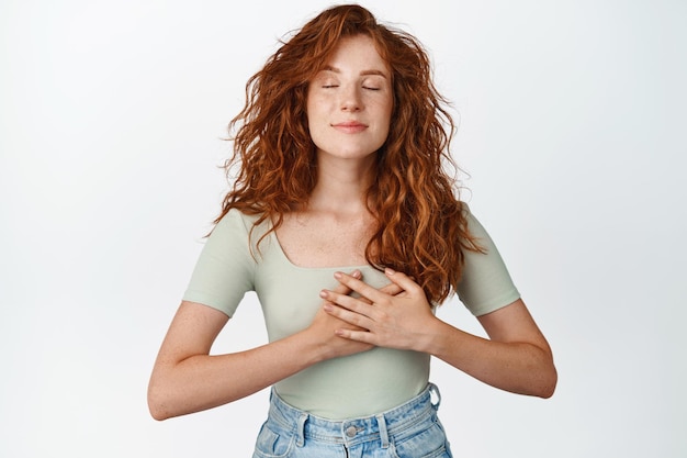 Free photo passionate redhead girl holding hands on heart close eyes and dreaming thinking of something heartwarming and dear to her standing over white background