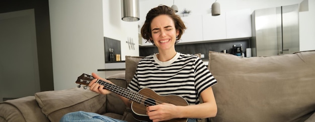 Free photo passionate young woman musician playing ukulele at home singing with joy sitting on couch in living