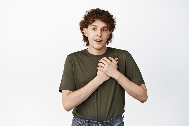 Passionated young man looking with admiration gazing fascinated at camera holding hands on heart standing over white background