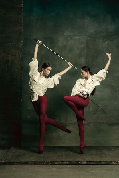 Free photo passioned. two young female ballet dancers like duelists with swords on dark green wall. caucasian models dancing together. ballet and contemporary choreography concept.