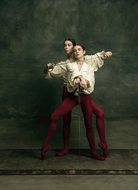 Passioned. Two young female ballet dancers like duelists with swords on dark green wall. Caucasian models dancing together. Ballet and contemporary choreography concept. 