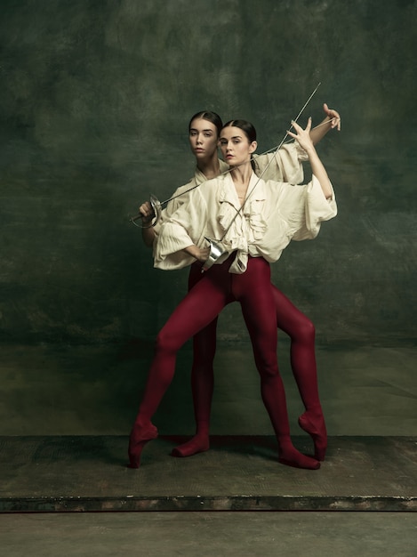 Passioned. Two young female ballet dancers like duelists with swords on dark green wall. Caucasian models dancing together. Ballet and contemporary choreography concept. 