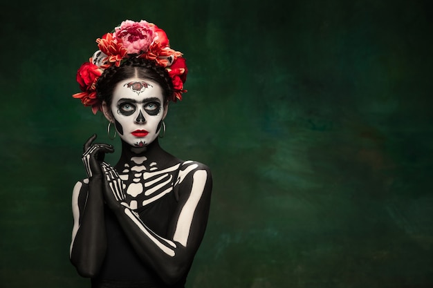 Passioned. young girl like santa muerte saint death or sugar skull with bright make-up. portrait isolated on dark green studio background with copyspace. celebrating halloween or day of the dead.