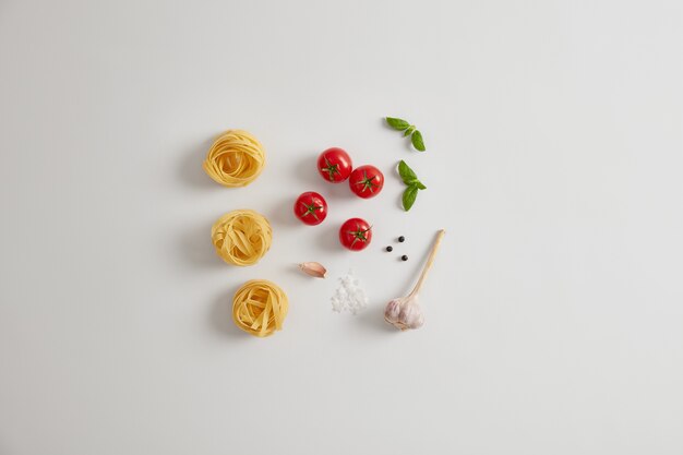 Pasta ingredients on white background. Red cherry tomatoes, basil, garlic, peppercorns, uncooked pasta nests for preparing tasty dish. Italian cuisine concept. Healthy vegetarian diet. Flat lay