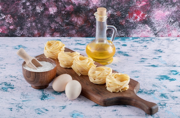 Pasta nests on wooden board with flour, eggs and olive oil.