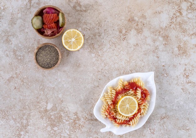 Pasta platter, lemon slice, assorted pickles and a bowl of black pepper on marble surface