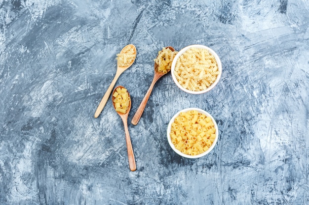 Pasta in white bowls and wooden spoons on a grungy plaster background. flat lay.