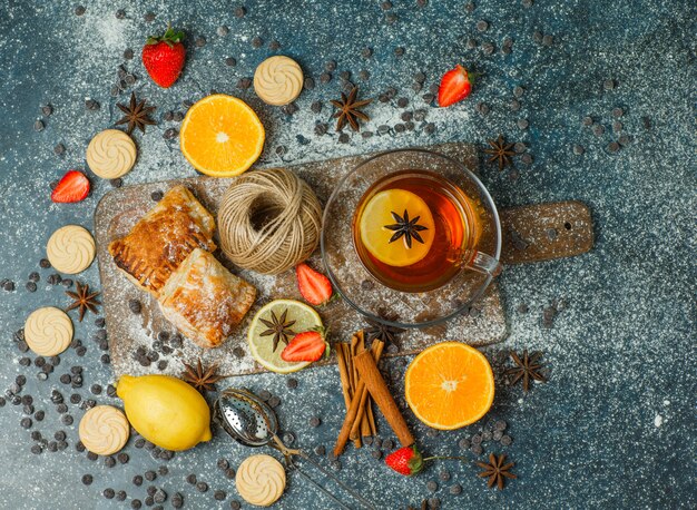 Pastries with flour, tea, fruits, cookies, choco chips, spices, thread on stucco and cutting board, top view.