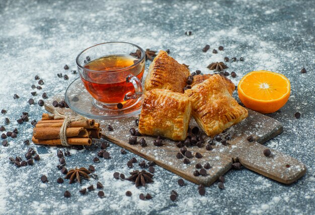 Pastries with tea, flour, choco chips, spices, orange on concrete and cutting board, high angle view.