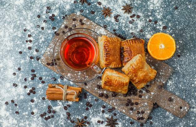 Pastries with tea, flour, choco chips, spices, orange on concrete and cutting board, top view.