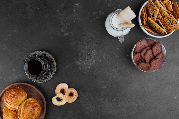 Pastry buns and crackers with a glass of tea and milk on black, top view.