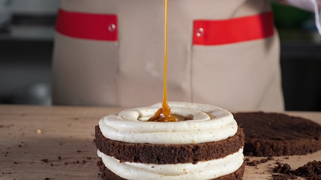 Pastry chef is pouring caramel on chocolate sponge cake with cream. baking and industrial food production. close-up view.