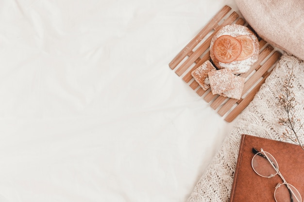 Pastry, eyeglasses, book and plaid lying on white bedsheet