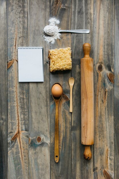 Кондитерские изделия; подвижный штифт; ложка; яйцо; мука и блокнот на деревянной поверхности