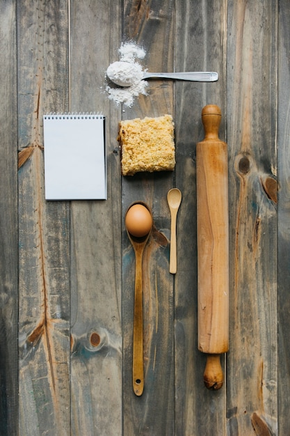 Pastry; rolling pin; spoon; egg; flour and notepad on wooden surface