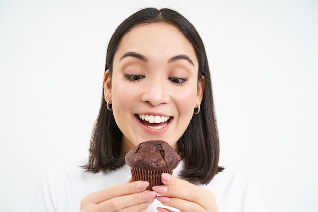 Free photo pastry sweets and bakery close up of smiling asian woman biting cupcake with joy standing isolated o