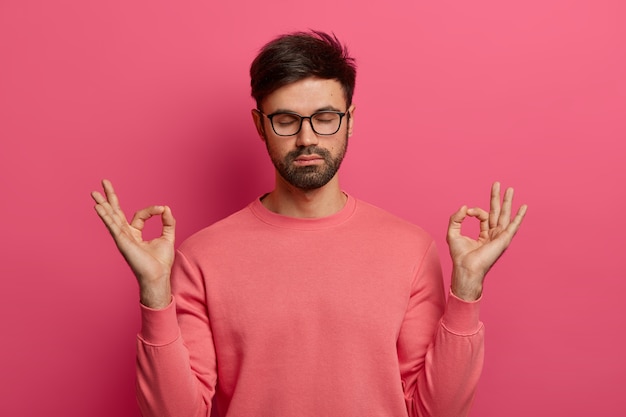 Free photo patience, calmness and meditation concept. peaceful relieved bearded young man practices yoga exercise, keeps hands in zen gesture, closes eyes, poses over pink wall, controls his emotions