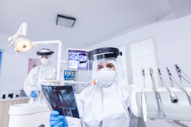 Patient first person view of dentist holding jaw radiography talking about tooth treatment. Dental specialist wearing protective hazmat suit against coroanvirus showing radiography.