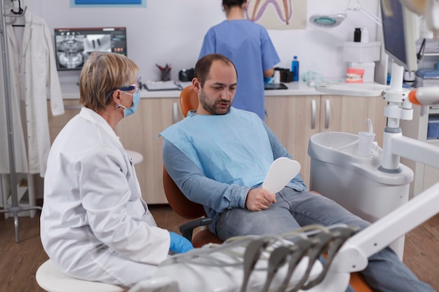 치과 진료실에서 치과 진료를 받는 동안 충치 감염 후 치아 절차를 보고 있는 거울을 들고 있는 환자. 구강 위생을 설명하는 치과 의사 수석 여자. 의학의 개념