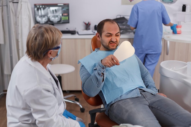 Пациент держит зеркало, глядя на процедуру зубов после стоматологической операции во время стоматологического осмотра в стоматологическом кабинете. Старшая женщина-дантист объясняет гигиену полости рта. Концепция медицины