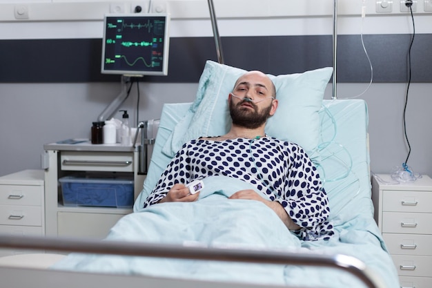 개인 병동에서 생체 측정 모니터에 연결된 호흡기 문제가 있는 병원 침대에 누워 있는 환자. 질병과 낮은 산소 포화도를 가진 중년 남성이 임상 상담을 기다리고 있습니다.