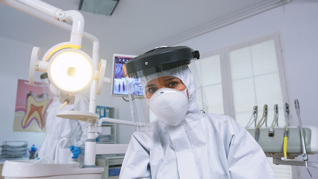 新しい通常の口腔病学オフィスでcovid防護服を着て歯の治療を説明する歯科医の患者のハメ撮り。患者のヘルスケアチェック中のコロナウイルスに対する安全装備のStomatolog。