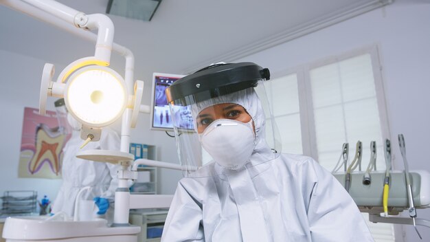 Patient pov of dentist explaining teeth treatment wearing covid protective suit in new normal stomatological office. Stomatolog in safety gear against coronavirus during heatlhcare check of patient.