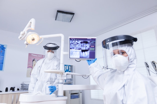 Пациент pov стоматолога с лицевой маской, объясняя стоматологический рентгеновский снимок, указывающий на монитор. Врач-стоматолог в защитном костюме от заражения коронавирусом указывает на рентгенографию.