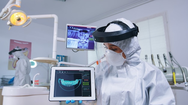 Foto gratuita spiegazione d'ascolto del punto di vista del paziente per il trattamento dei denti, dentista in tuta che mostra i raggi x sul tablet. specialista in stomatologia che indossa tuta protettiva contro l'infezione da covid19 che punta alla radiografia