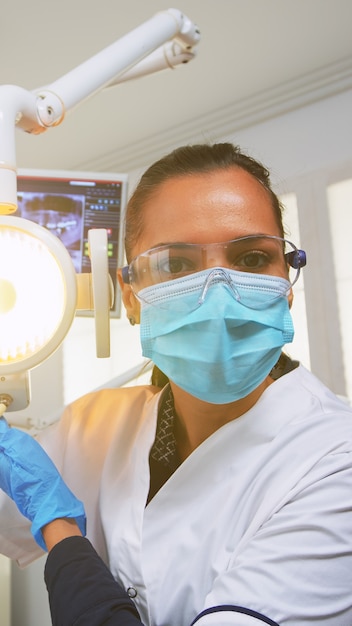 환자 pov는 영향을받는 질량을 치료하는 수술을 위해 치과 진료소를 방문합니다. 현대 교정 사무실에서 함께 일하는 의사와 간호사, 램프를 밝히고 보호 마스크를 쓴 사람을 검사합니다.