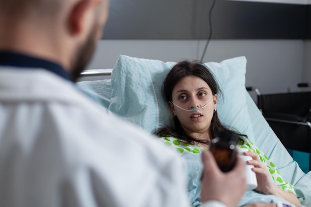 病院のベッドで回復している処方薬のボトルを提示している医師を見ている酸素を受け取っている鼻カニューレの患者。手術後に女性に鎮痛剤を与える薬剤師の医者。