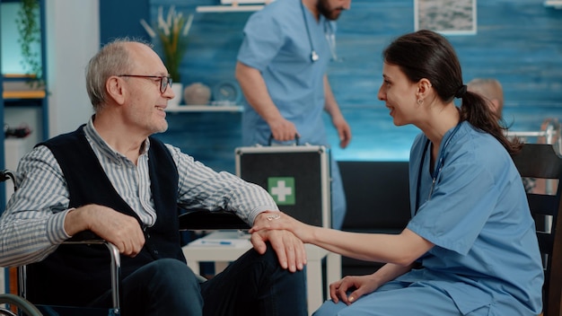 Patient with physical disease getting assistance from nurse in nursing home. Medical assistant talking to elder man sitting in wheelchair about healthcare treatment and recovery therapy.