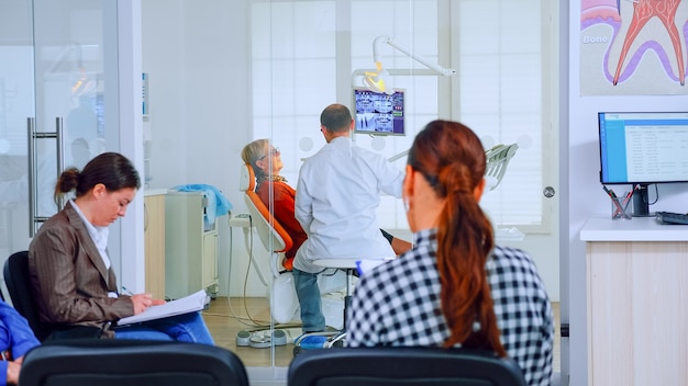 Patients sitting on chairs in waiting room of stomatological clinic filling in stomatological forms while doctor working in background. Concept of crowded professional orthodontist reception office.