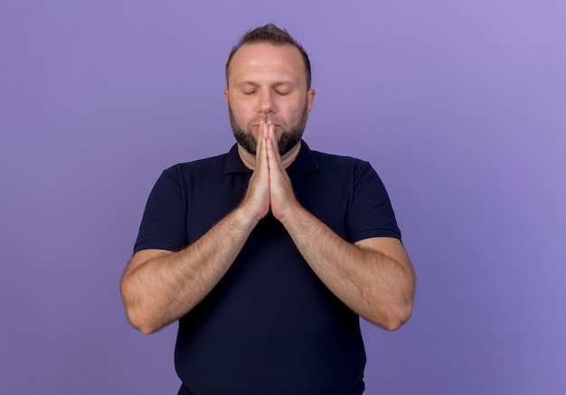 Мирный взрослый славянский мужчина, складывая руки возле рта, молится с закрытыми глазами изолированы