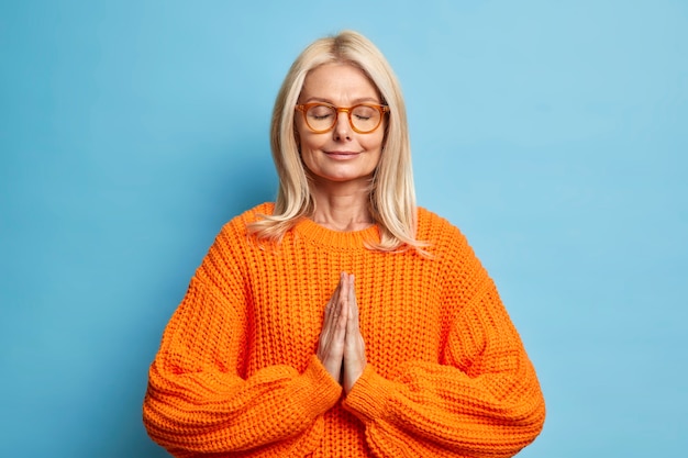 Peaceful blonde woman keeps eyes closed stands in praying pose prays for good wellness dressed in knitted jumper wears spectacles.