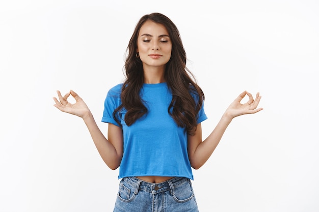 Foto gratuita tranquilla e calma bella donna bruna in t-shirt blu, fare un gesto zen, in piedi posa del loto, respirare calmo e rilassato, inspirare aria durante la meditazione, muro bianco