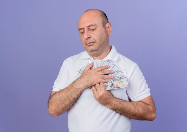 Peaceful casual mature businessman holding money with closed eyes isolated on purple background with copy space