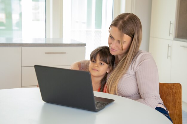 Peaceful girl and her mom using laptop, sitting at table and hugging, watching movie, looking at display.