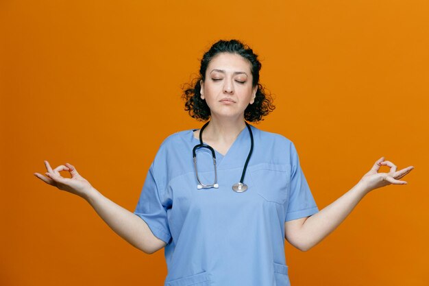 Peaceful middleaged female doctor wearing uniform and stethoscope around her neck meditating with closed eyes isolated on orange background